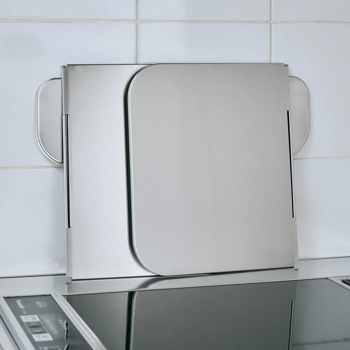 Foldable stainless steel stove splatter guard folded compactly and leaning against a tiled kitchen wall