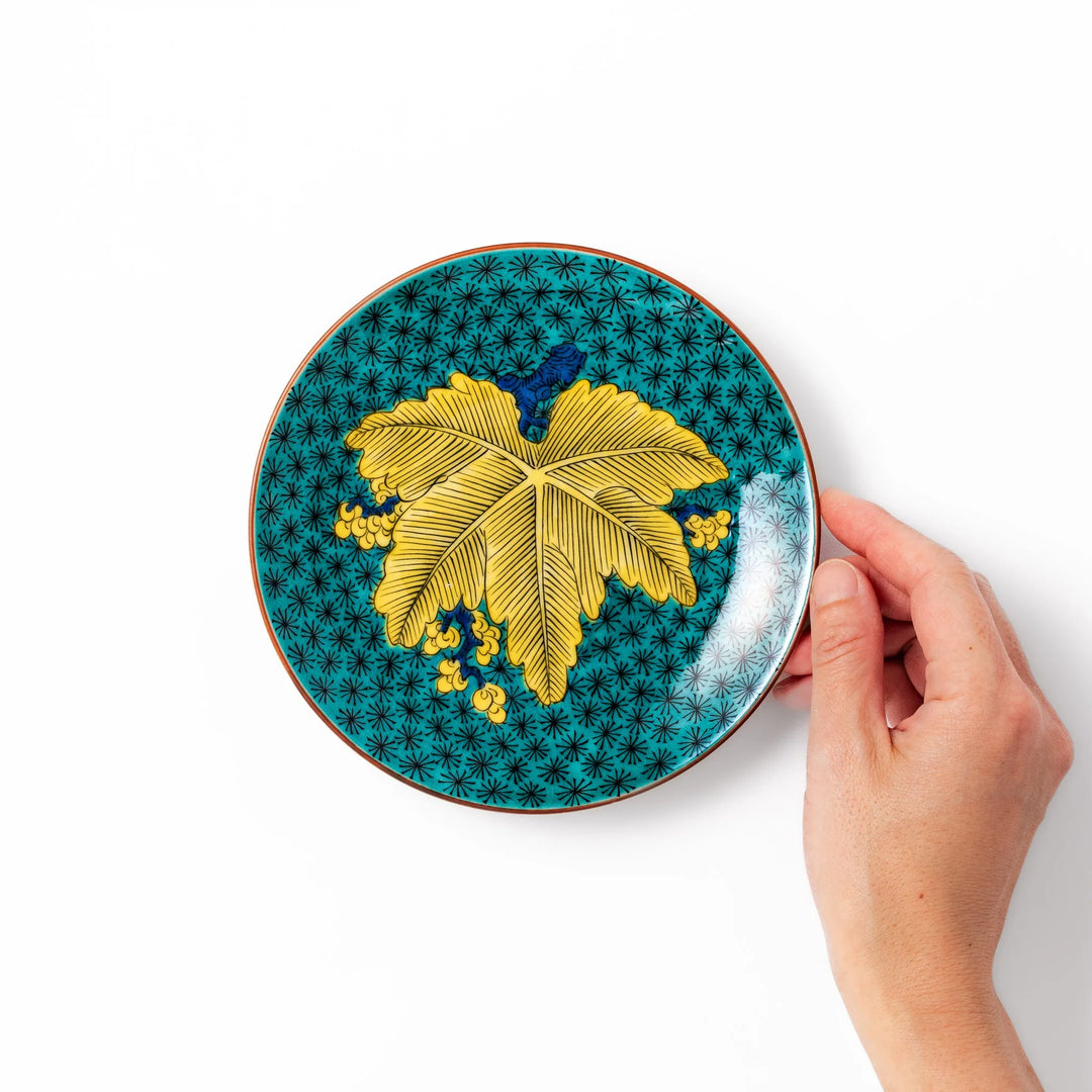 Round Kutani dessert plate with a bold golden leaf and grape motif on a turquoise background patterned with black starbursts.