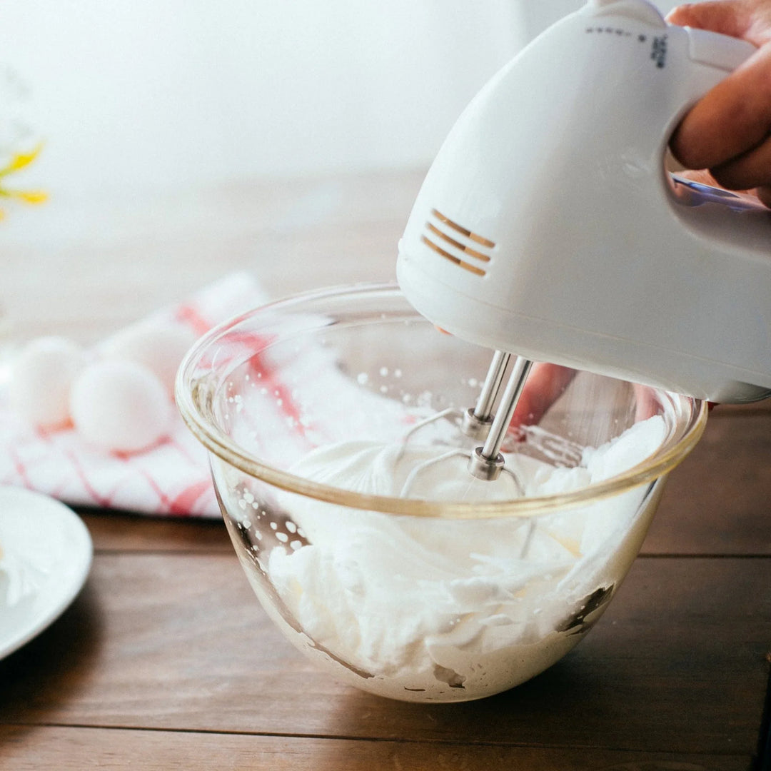 Whipping white cream with electric hand mixer in hario heatproof glass bowl on wooden table baking preparation