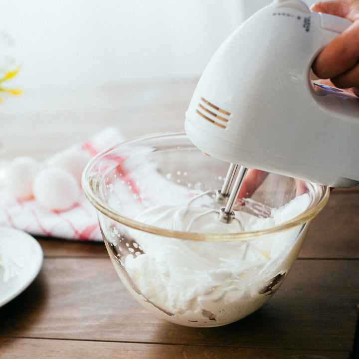 Whipping white cream with electric hand mixer in hario heatproof glass bowl on wooden table baking preparation