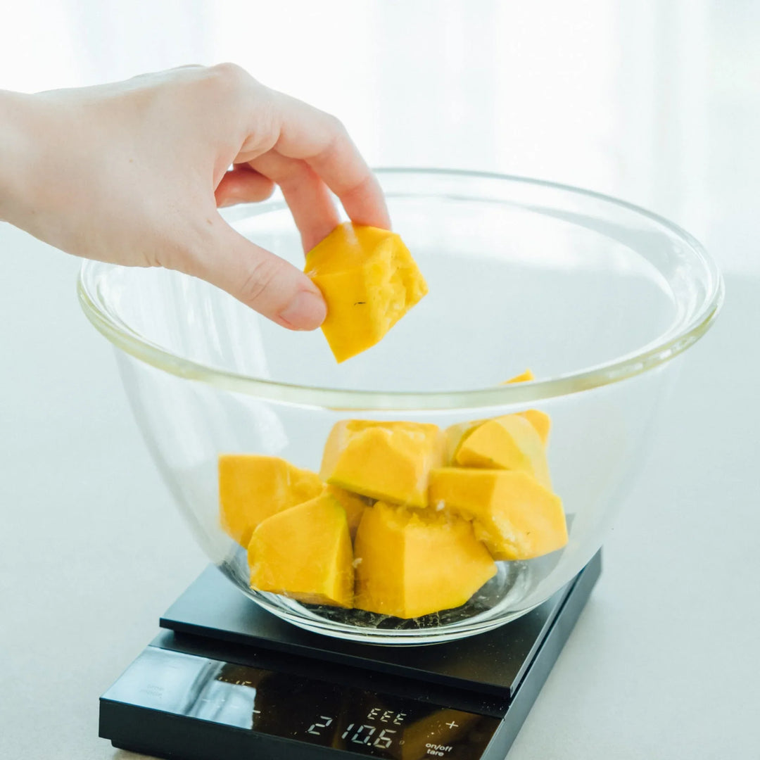 Placing chopped yellow pumpkin into hario heatproof glass mixing bowl on black digital food scale kitchen prep