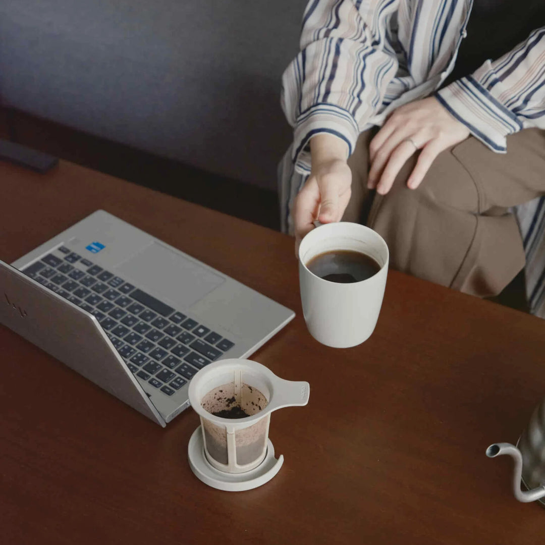 Holding hario ceramic tea and coffee mug filled with fresh brew next to mesh filter resting on lid by a laptop