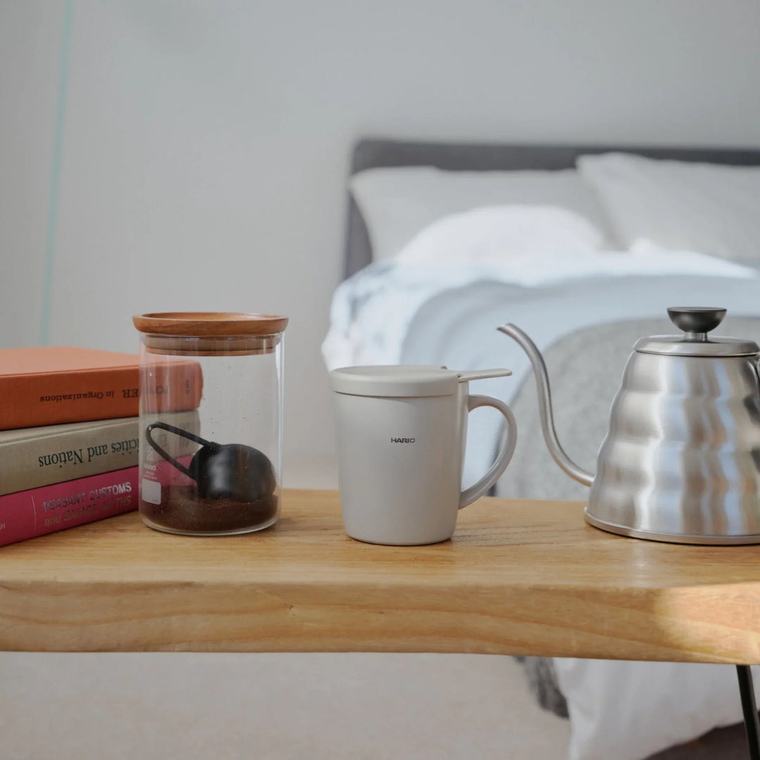 Hario ceramic tea and coffee mug with lid standing on wooden bedside table next to steel kettle and glass jar