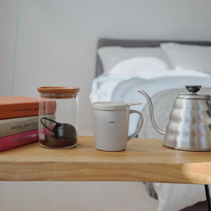 Hario ceramic tea and coffee mug with lid standing on wooden bedside table next to steel kettle and glass jar