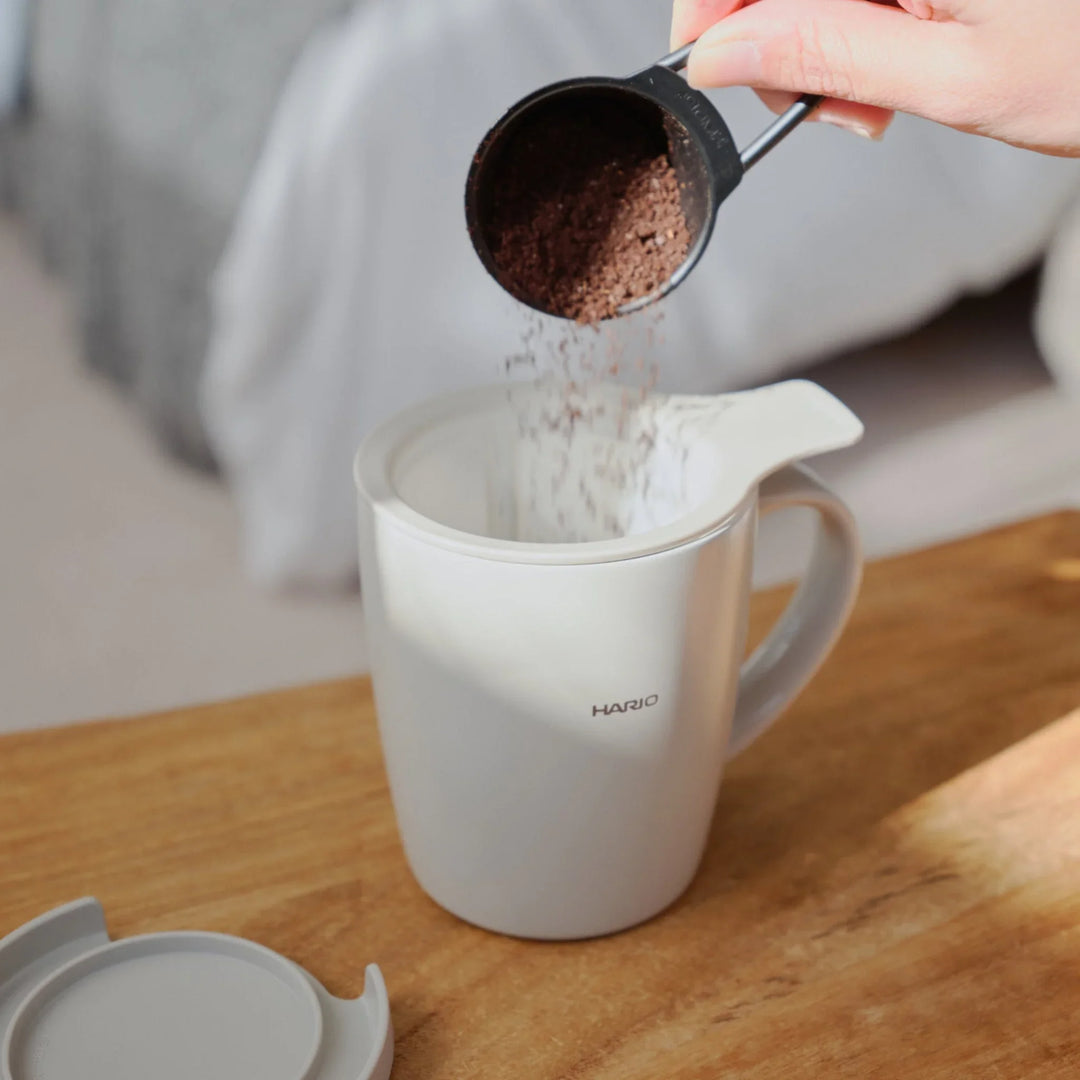 Pouring coffee grounds from a scoop into mesh filter inside hario ceramic tea and coffee mug on wooden table