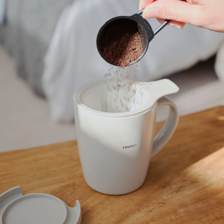 Pouring coffee grounds from a scoop into mesh filter inside hario ceramic tea and coffee mug on wooden table