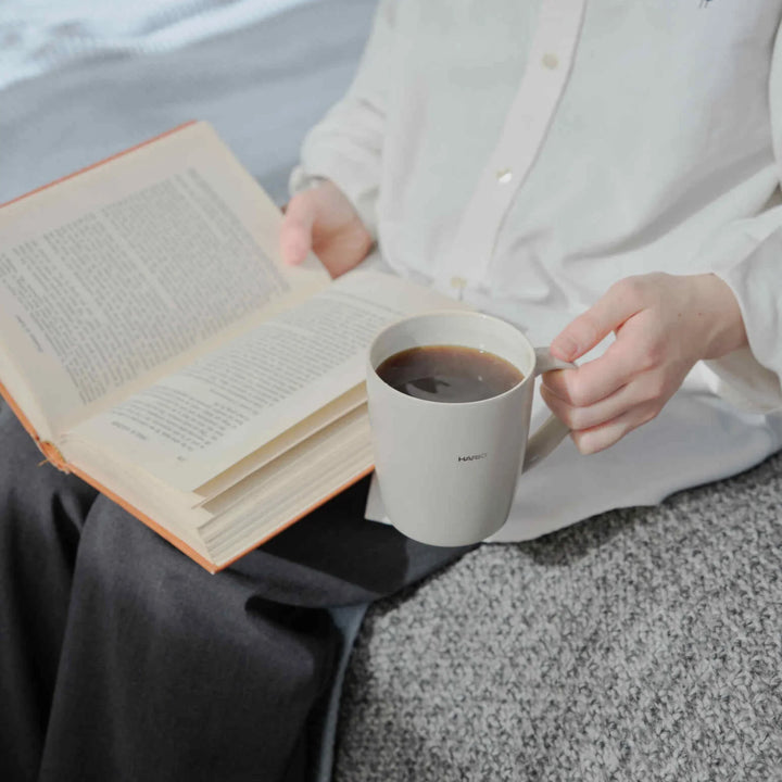 Holding hario ceramic tea and coffee mug while reading an open book in bed during a relaxing morning routine