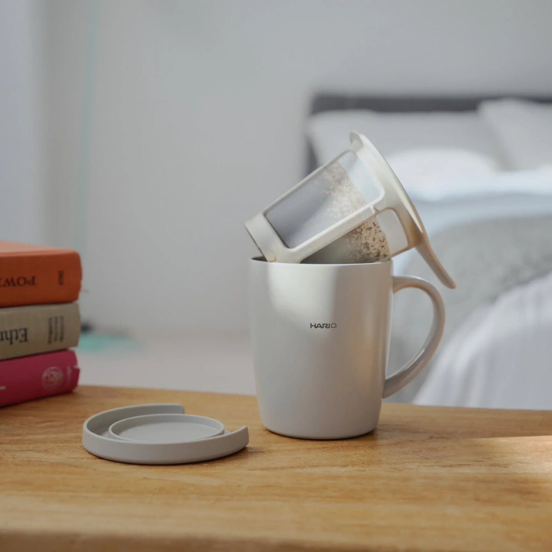 Pouring hot water from kettle into hario ceramic tea and coffee mug with infuser filter on wooden bedroom table