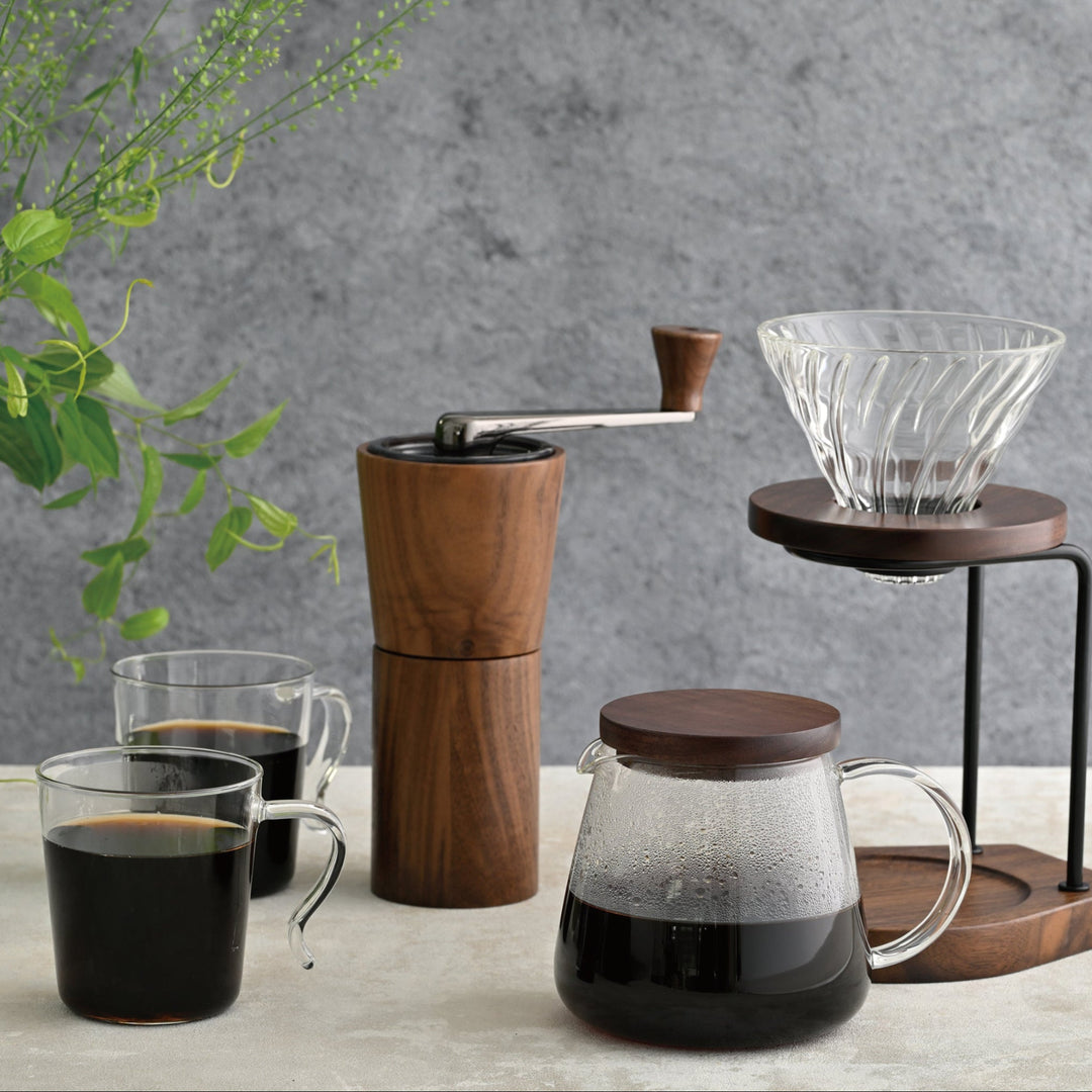 Displaying hario walnut wood coffee grinder on counter with glass mugs of brewed coffee and pour over dripper stand