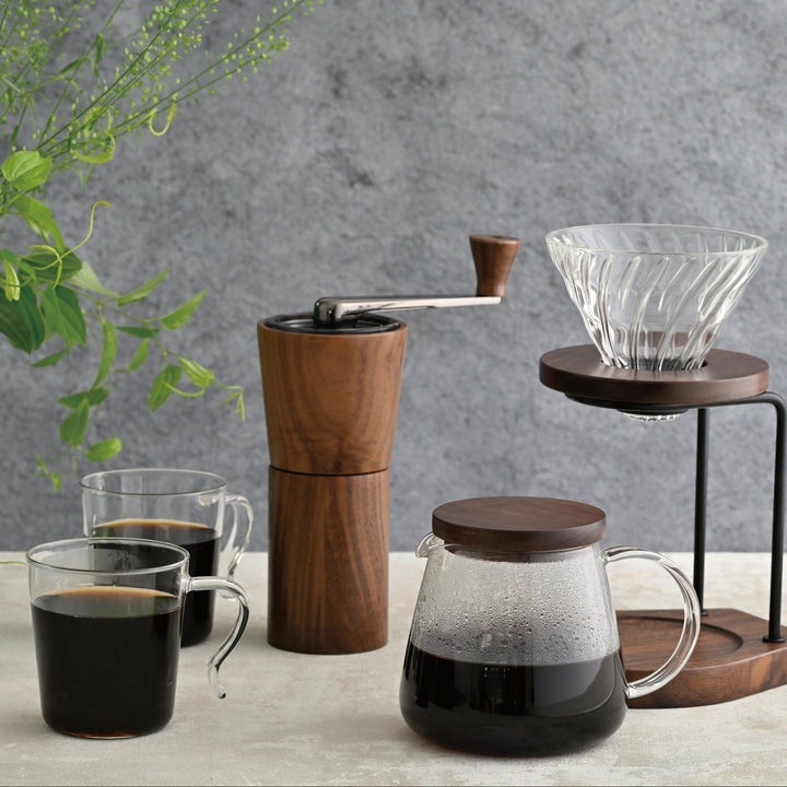 Displaying hario walnut wood coffee grinder on counter with glass mugs of brewed coffee and pour over dripper stand