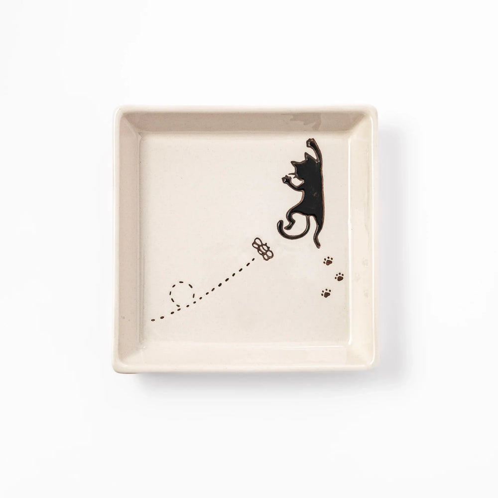 Square beige plate featuring a playful black cat chasing a butterfly, with paw prints and a dotted flight path.