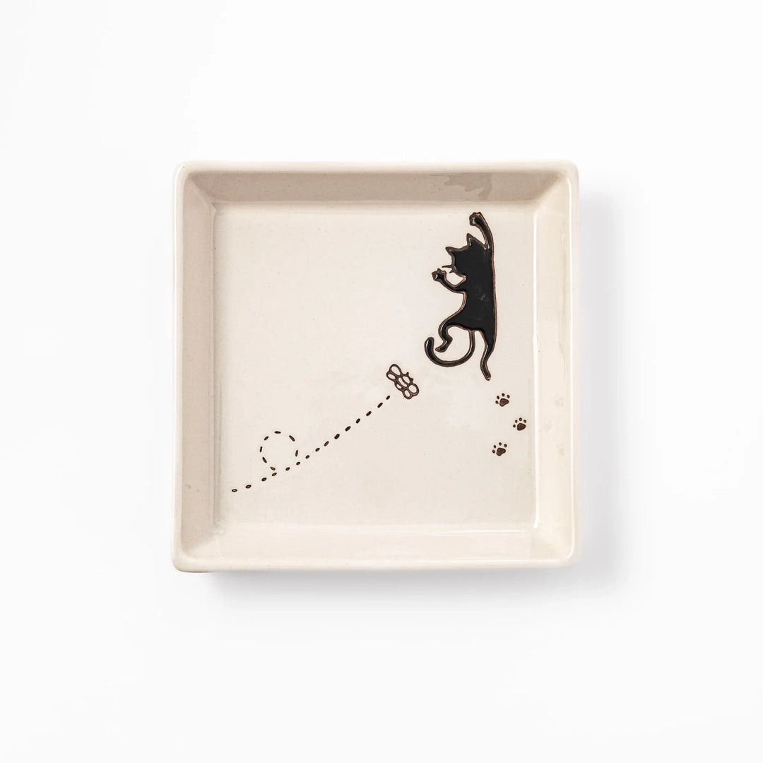 Square beige plate featuring a playful black cat chasing a butterfly, with paw prints and a dotted flight path.