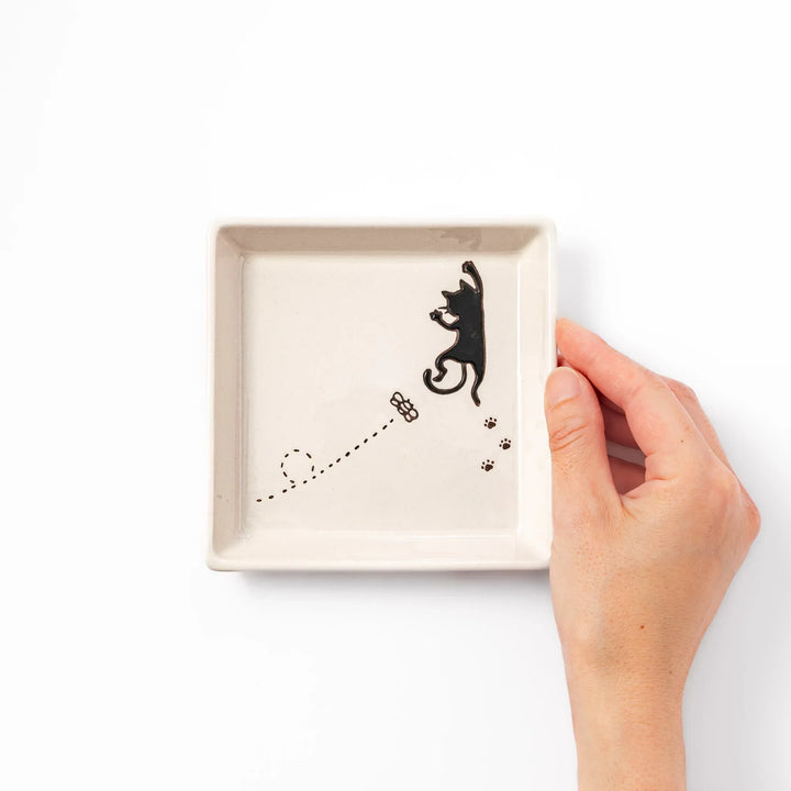 Square beige plate featuring a playful black cat chasing a butterfly, with paw prints and a dotted flight path.