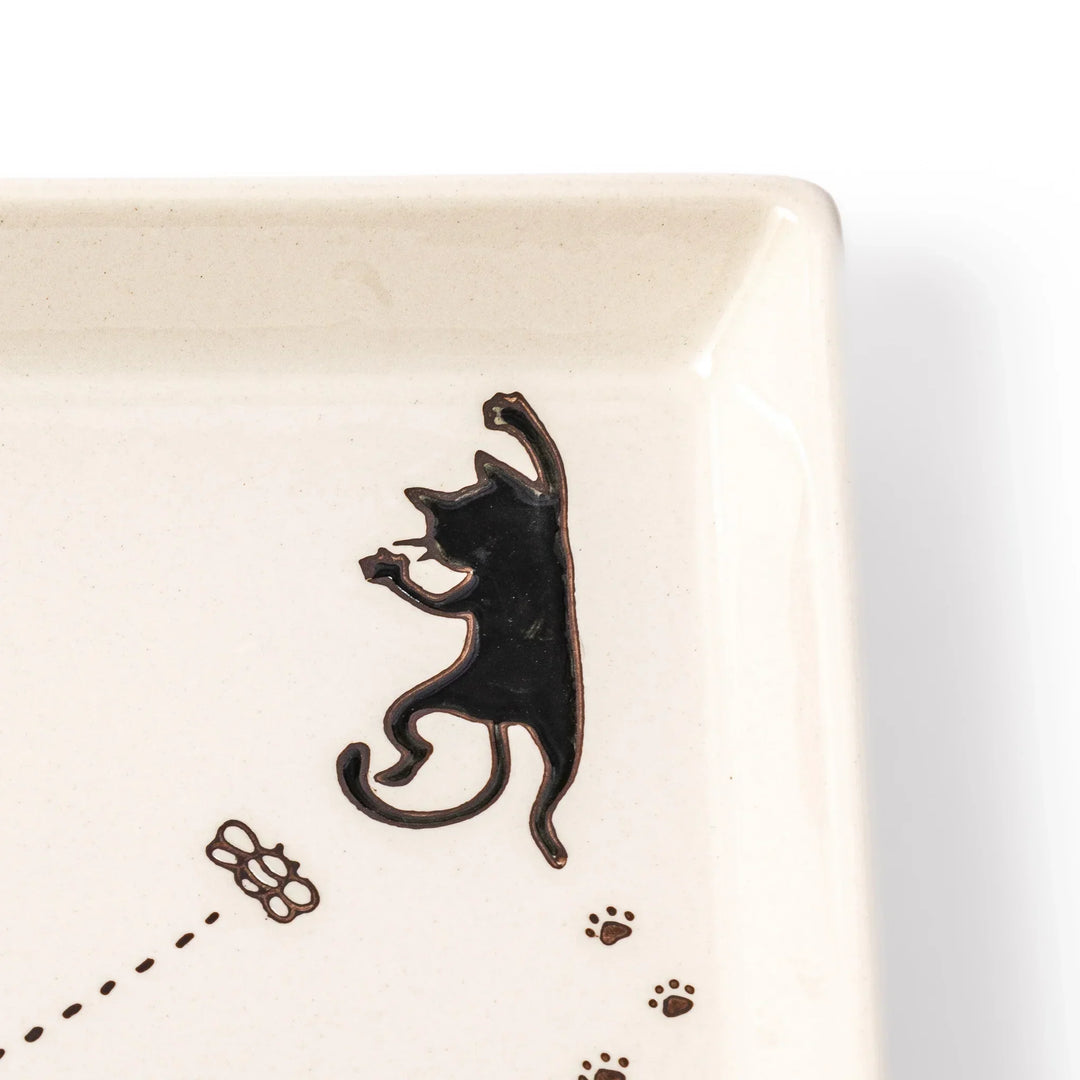 Square beige plate featuring a playful black cat chasing a butterfly, with paw prints and a dotted flight path.