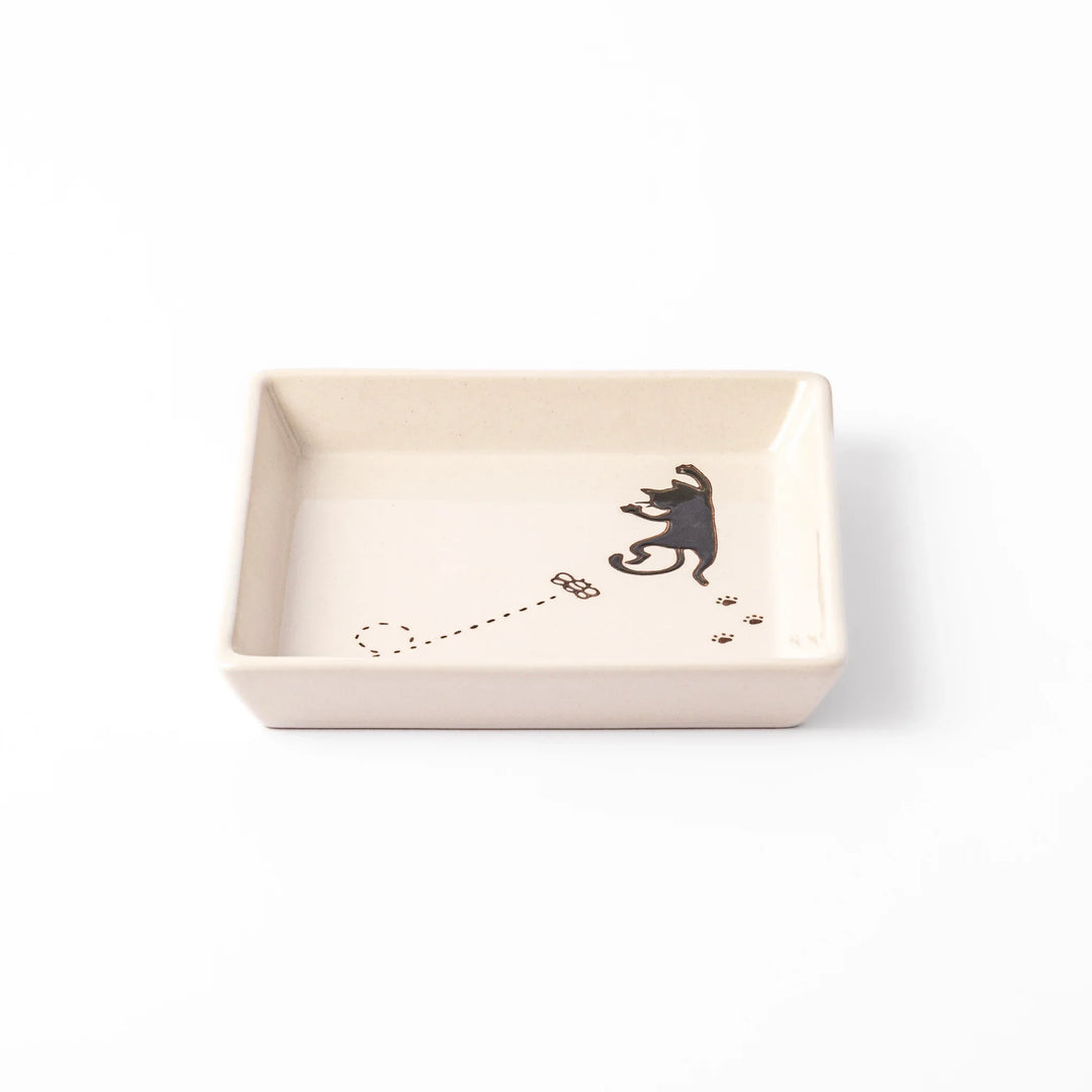 Square beige plate featuring a playful black cat chasing a butterfly, with paw prints and a dotted flight path.