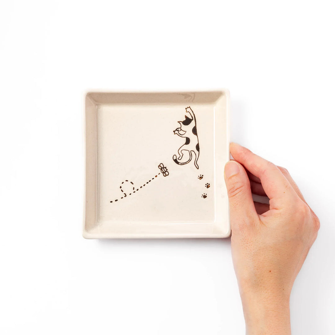 Beige square plate with playful calico cat and butterfly illustration, accented with dotted lines and paw prints.