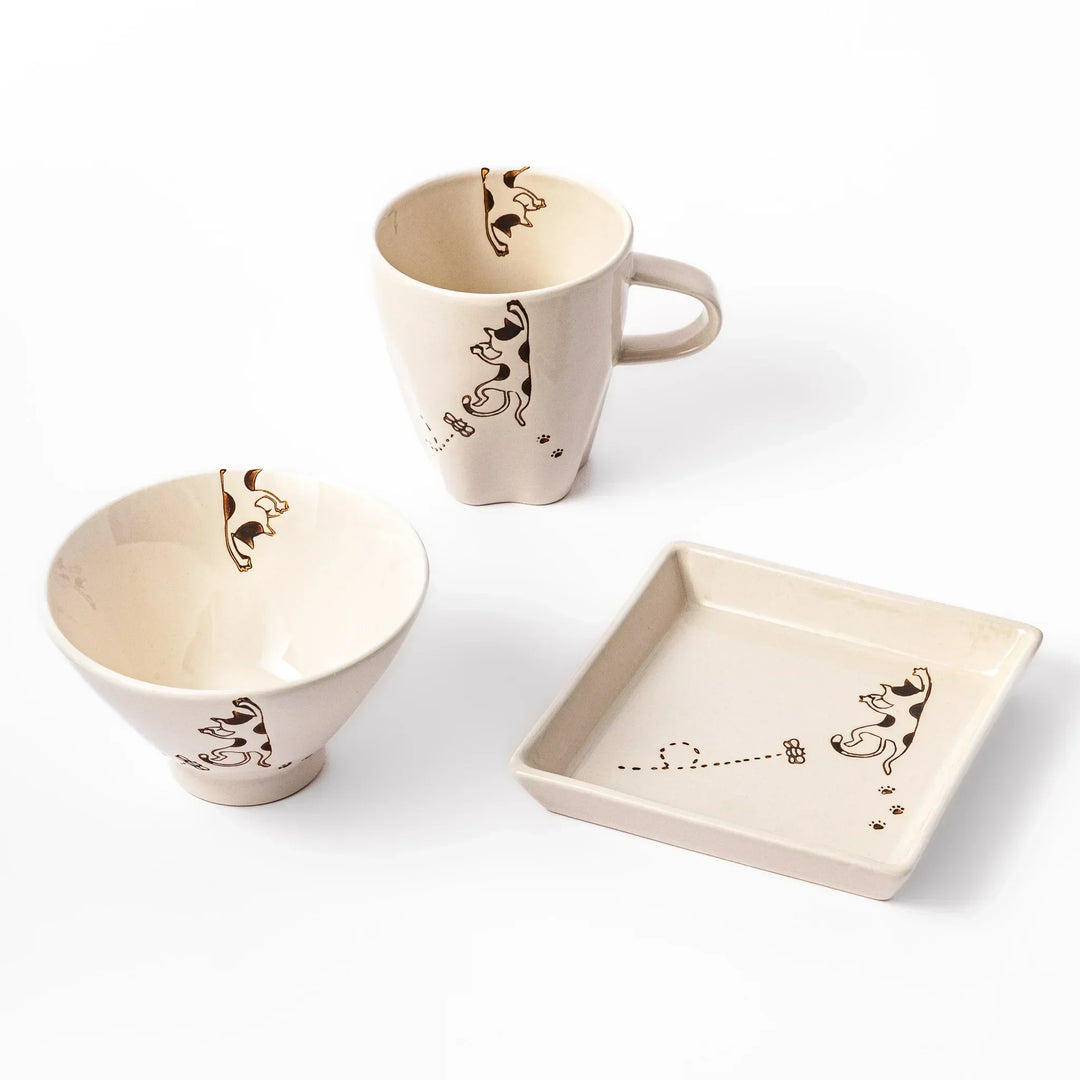 Beige mug, bowl, and square plate featuring calico cat and butterfly motif with paw prints and playful dotted lines.