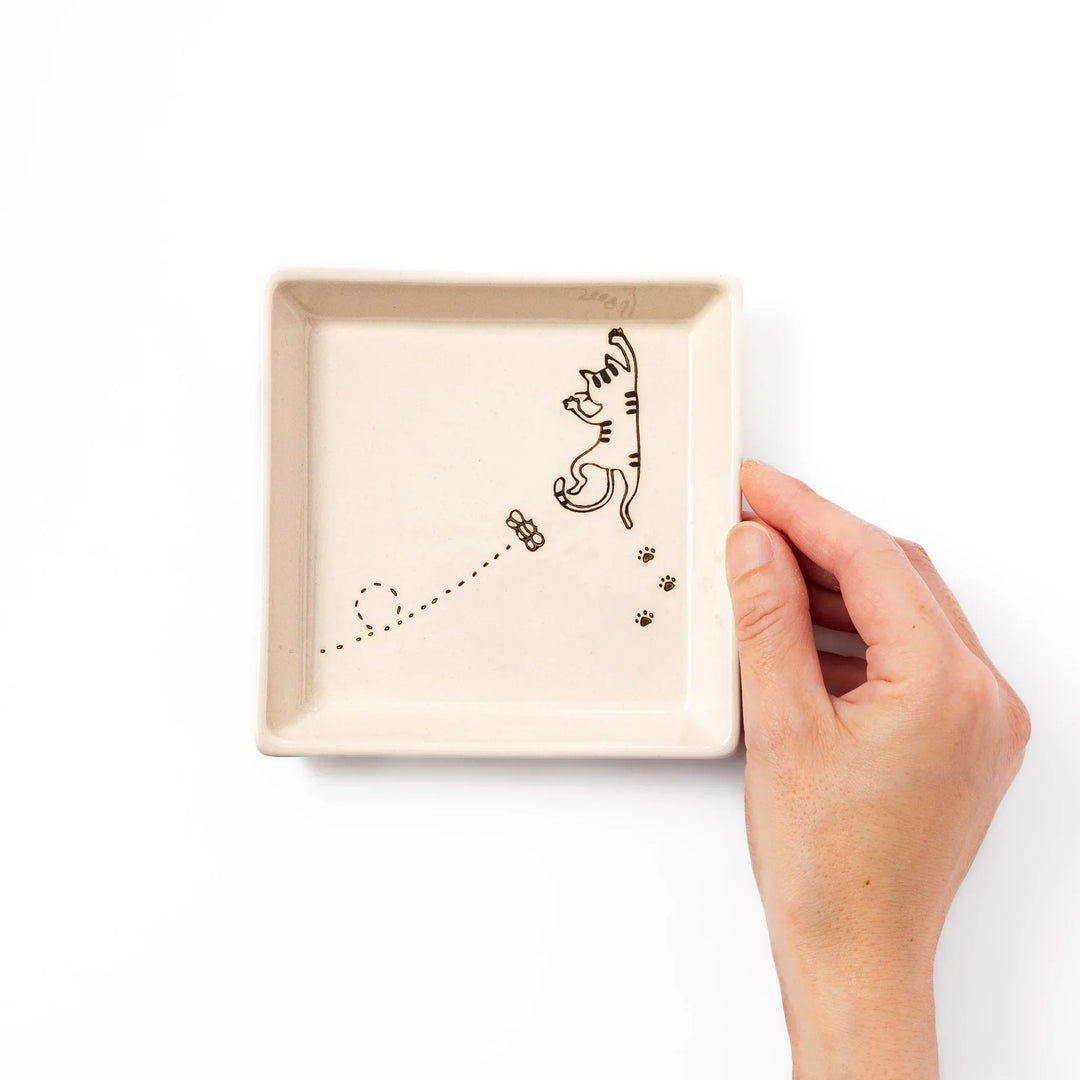 Beige square plate featuring a tabby cat chasing a butterfly with dotted trail and small paw prints.