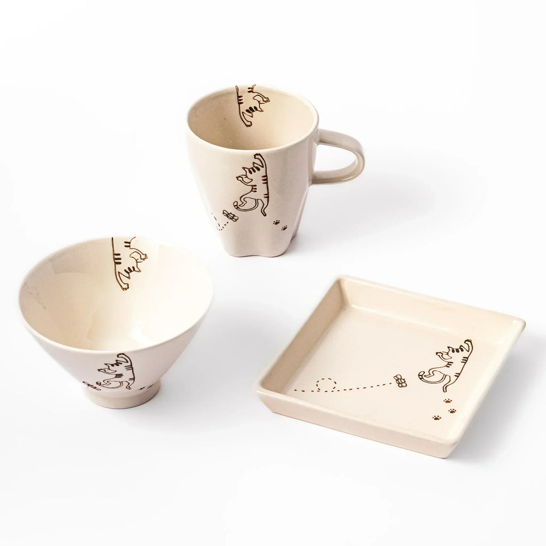 Matching mug, bowl, and square plate featuring a striped cat chasing a butterfly with dotted lines and paw prints.