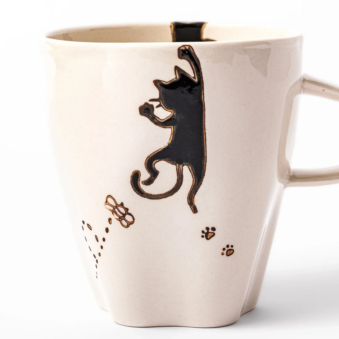 Cream-colored cup with a black cat design reaching for a butterfly, accented by playful dotted lines and paw prints.