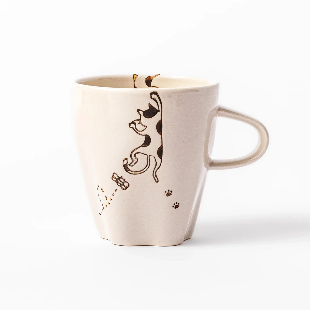 Cream-colored cup with a calico cat design reaching for a butterfly, decorated with dotted lines and paw prints.