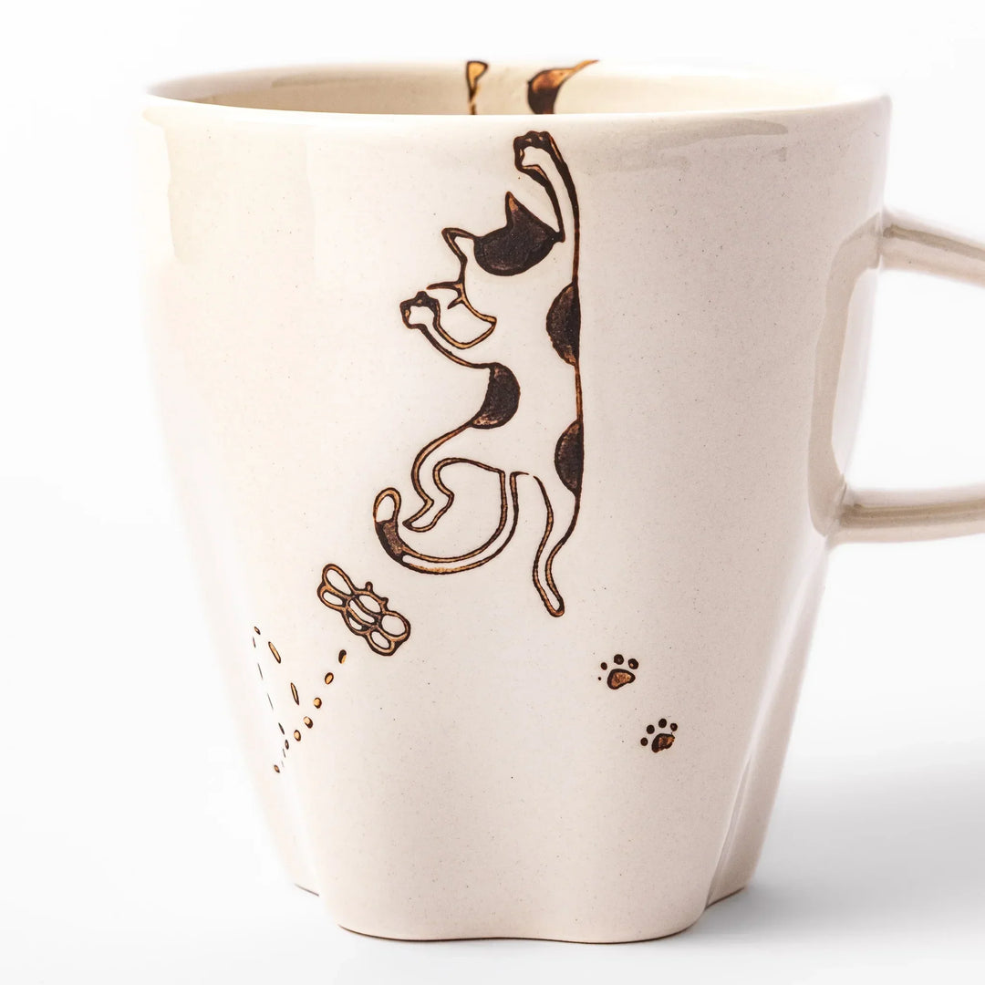 Cream-colored cup with a calico cat design reaching for a butterfly, decorated with dotted lines and paw prints.