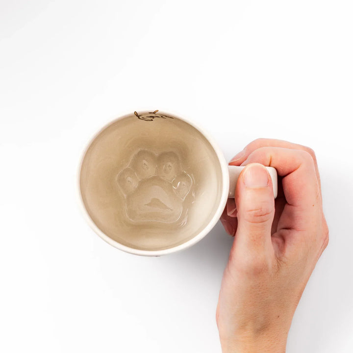 Cream-colored cup with a striped cat design reaching toward a butterfly, with dotted lines and paw prints.