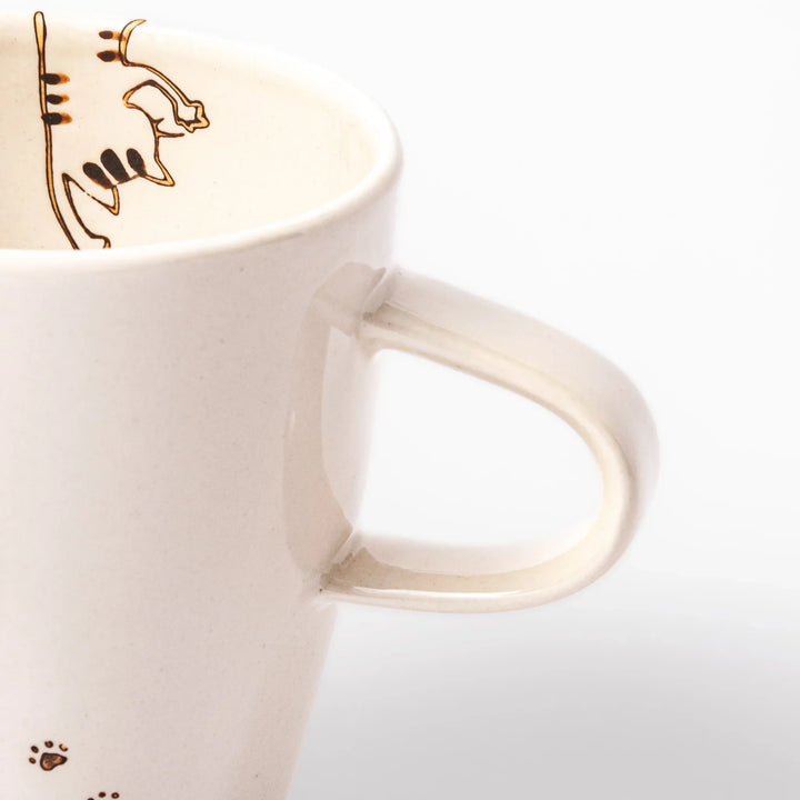 Cream-colored cup with a striped cat design reaching toward a butterfly, with dotted lines and paw prints.