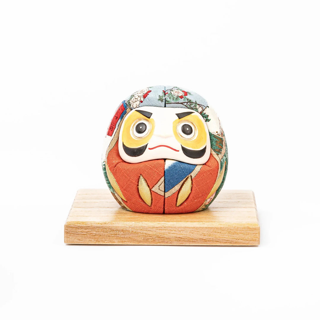 Fabric daruma ornament with bright multicolor kimono pattern and bold painted face displayed on a wooden stand