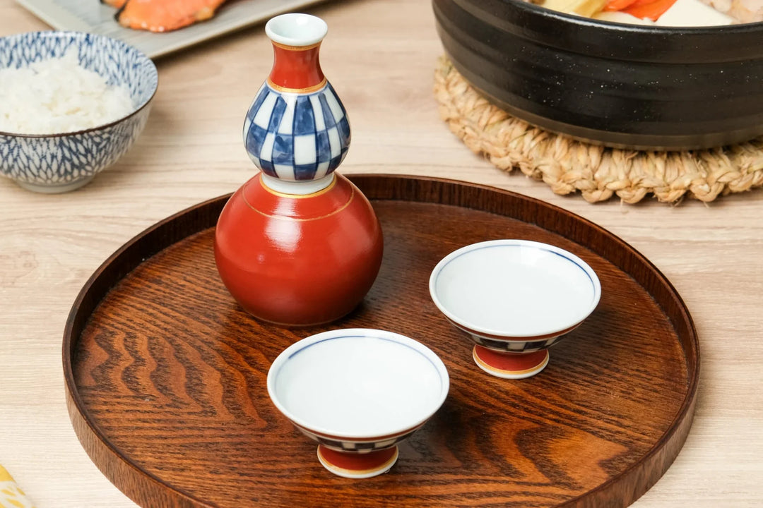 Japanese sake set with red and blue porcelain tokkuri and cups on a round wooden tray with a meal setting in the background