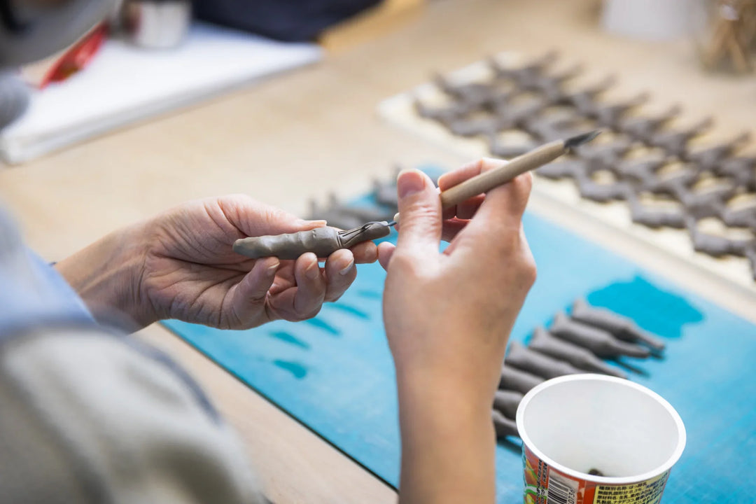Clay artisan shaping a small figurine by hand, using a carving tool with rows of unfinished pieces drying on the table.