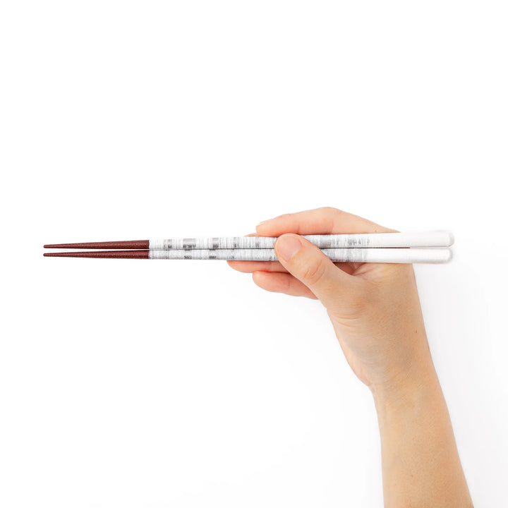 A hand holding a pair of silver textured Japanese chopsticks showcasing the elegant white and metallic design