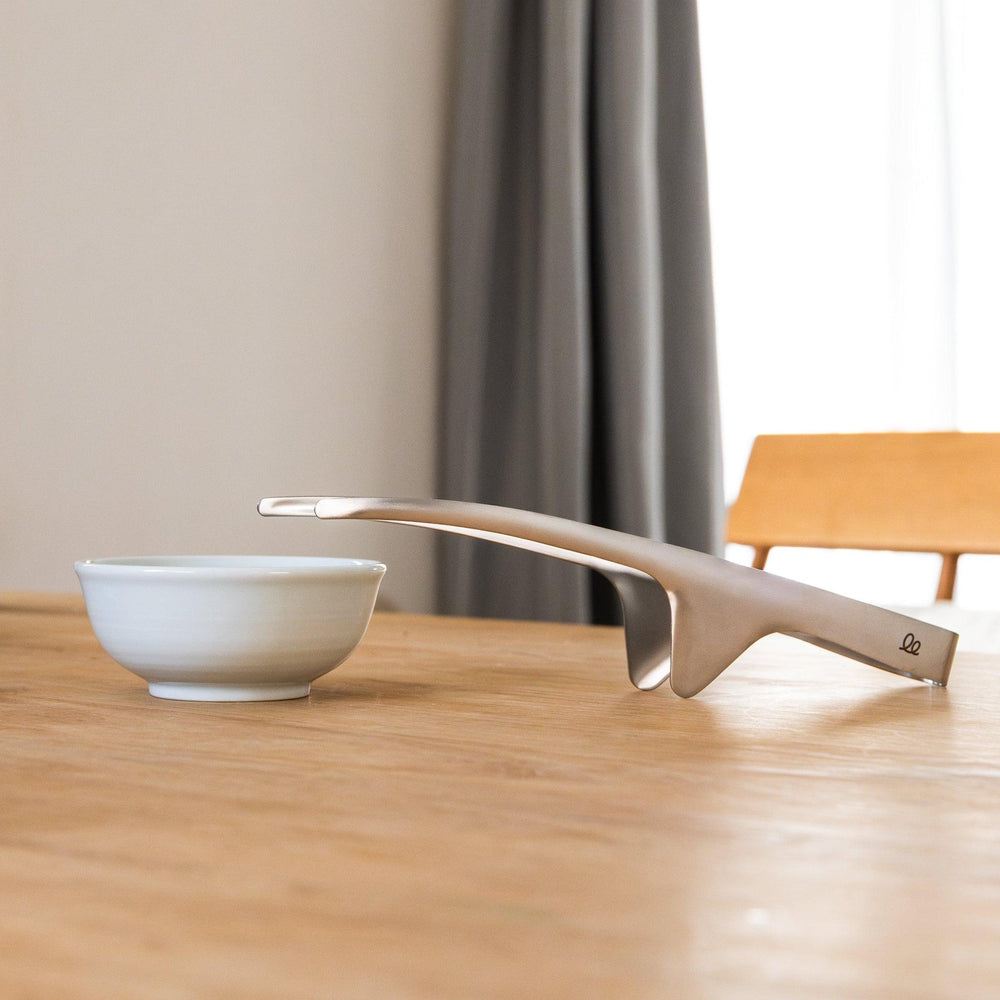 Japanese stainless tabletop BBQ tongs resting beside small white bowl on dining table
