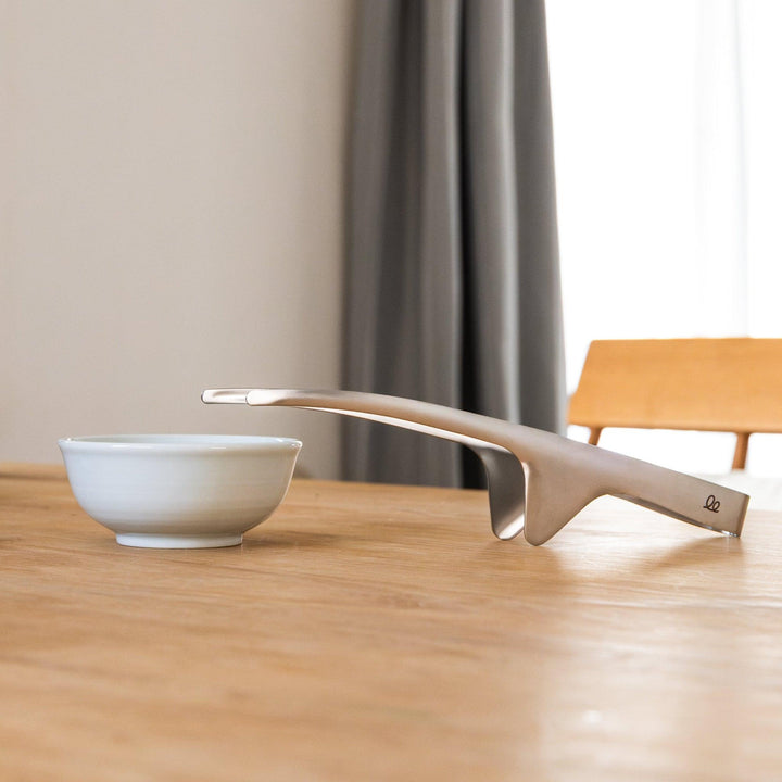 Japanese stainless tabletop BBQ tongs resting beside small white bowl on dining table