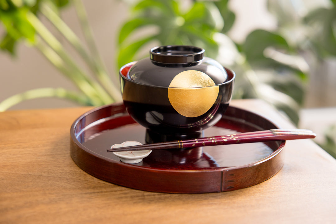 Black lacquer miso soup bowl with a gold moon design placed on a glossy round tray with chopsticks in a warm dining setting