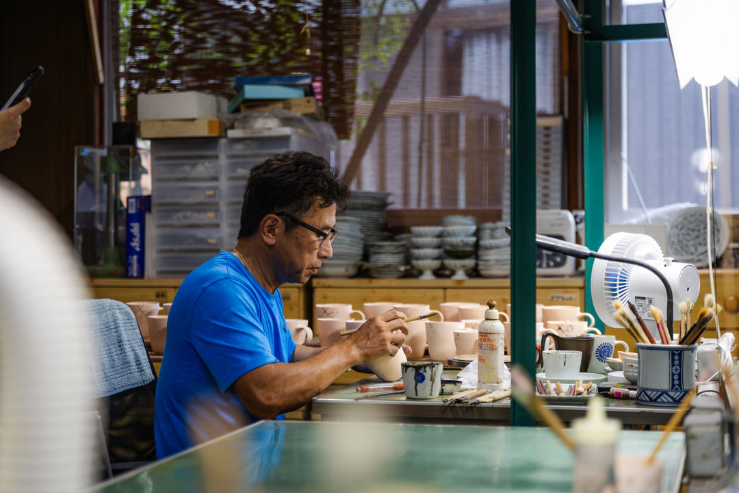 An artisan in a blue shirt hand-painting delicate designs onto a bisque-fired ceramic cup at a studio worktable.