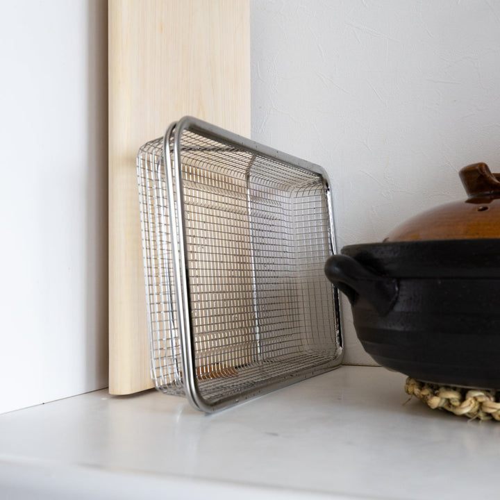 Compact stainless steel draining basket stored upright on a kitchen counter next to a Donabe and cutting board to save space