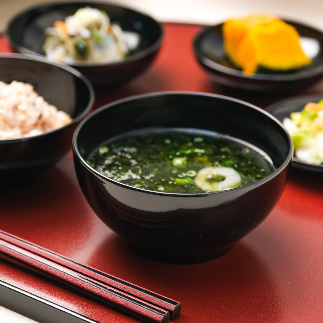 Black lacquer soup bowl with seaweed soup on red Japanese tray alongside rice and seasonal side dishes