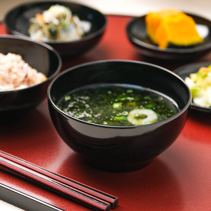 Black lacquer soup bowl with seaweed soup on red Japanese tray alongside rice and seasonal side dishes
