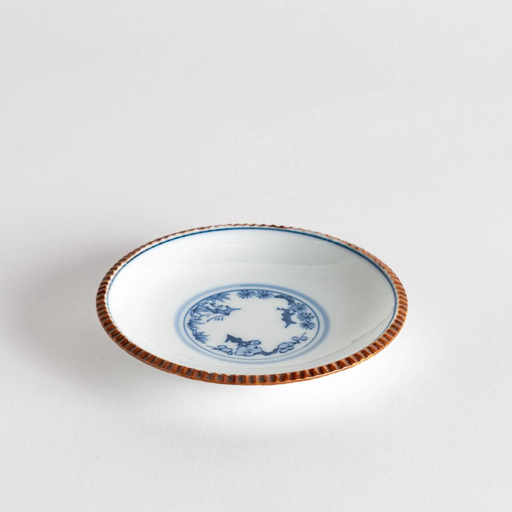 Small ceramic plate with central blue floral motif on white background framed by copper textured rim.
