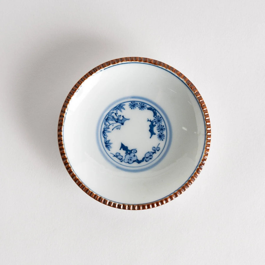 Small ceramic plate with central blue floral motif on white background framed by copper textured rim.