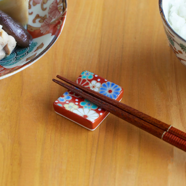 Japanese chopstick rest with red background and plum and chrysanthemum blossoms, holding wooden chopsticks on a wooden table.