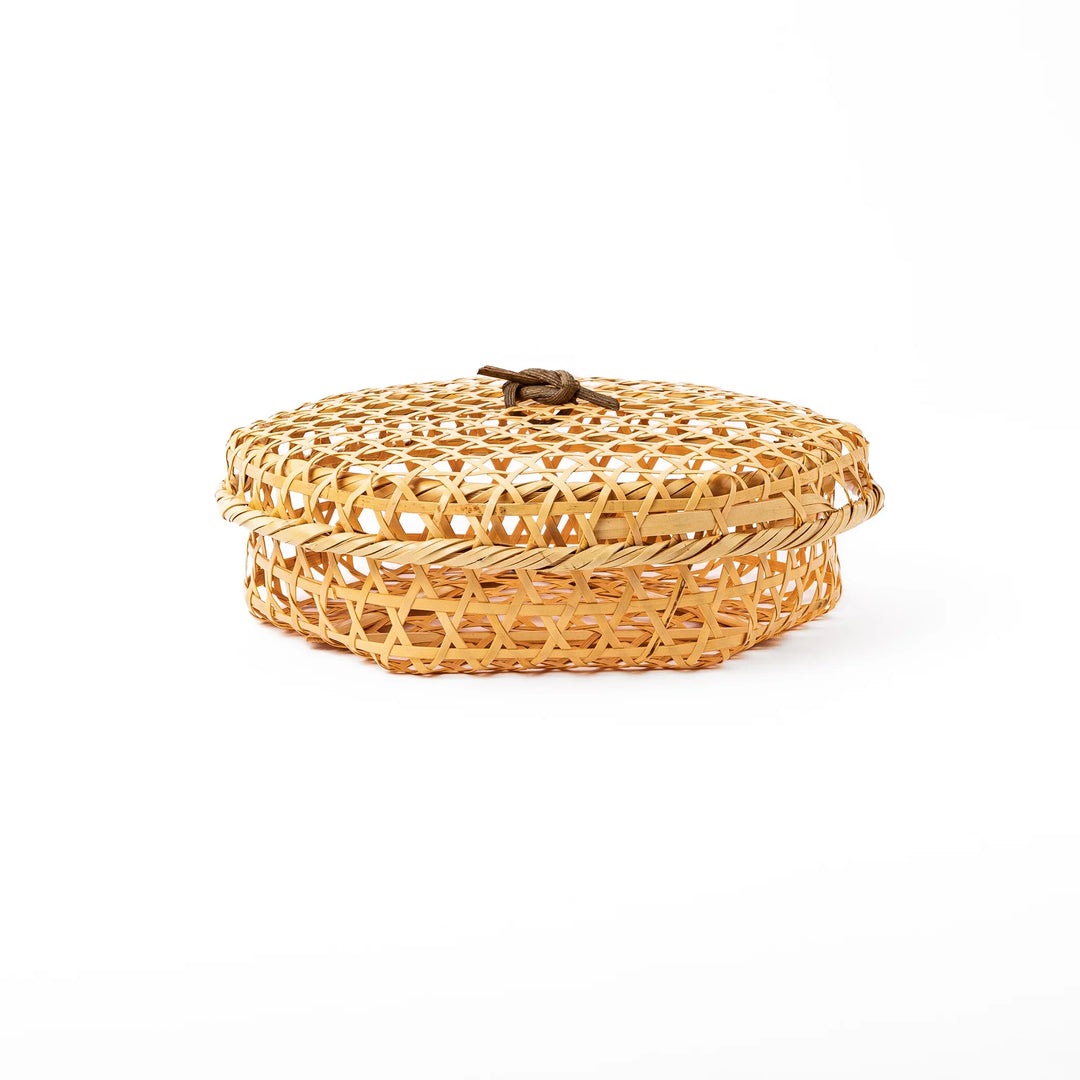 A closed round handwoven bamboo serving basket featuring a decorative brown knotted cord handle on top of the lid
