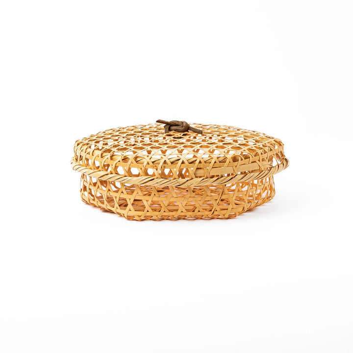 A closed round handwoven bamboo serving basket featuring a decorative brown knotted cord handle on top of the lid