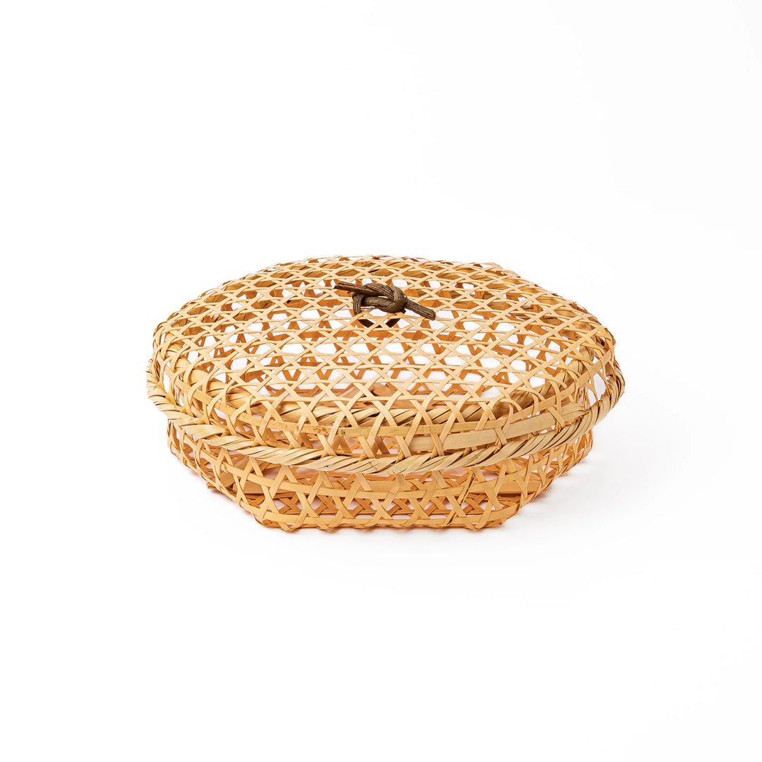 A closed round handwoven bamboo serving basket featuring a decorative brown knotted cord handle on top of the lid