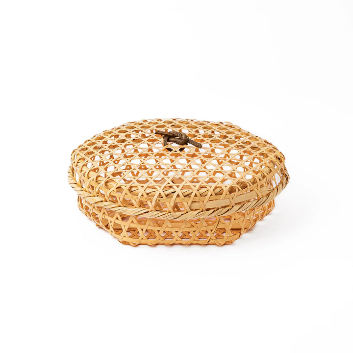 A closed round handwoven bamboo serving basket featuring a decorative brown knotted cord handle on top of the lid