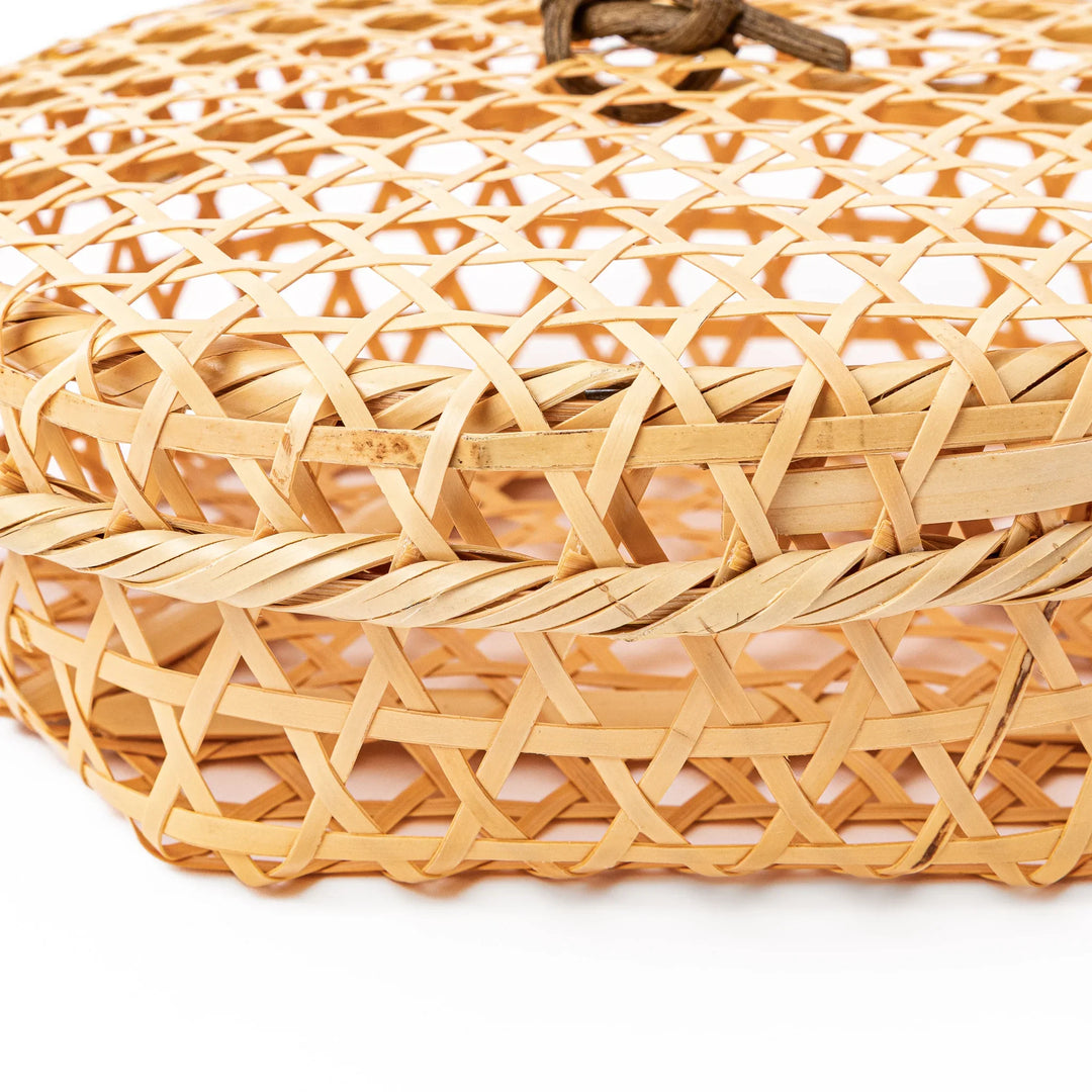 A closed round handwoven bamboo serving basket featuring a decorative brown knotted cord handle on top of the lid