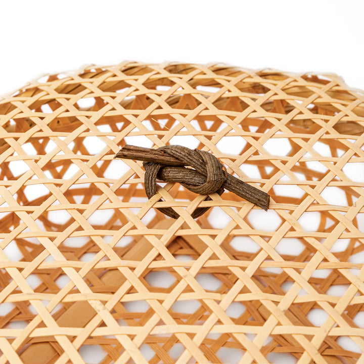 A closed round handwoven bamboo serving basket featuring a decorative brown knotted cord handle on top of the lid