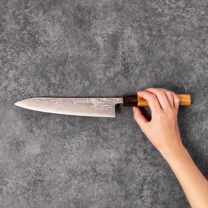 Japanese Damascus gyuto knife featuring a layered steel blade and natural wood handle displayed on gray stone background.