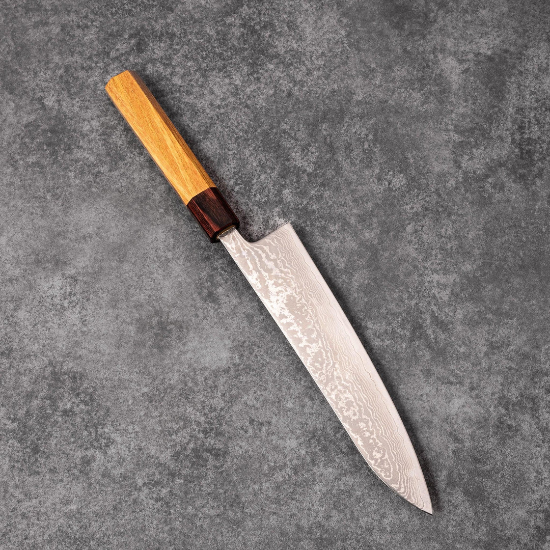 Japanese Damascus gyuto knife featuring a layered steel blade and natural wood handle displayed on gray stone background.